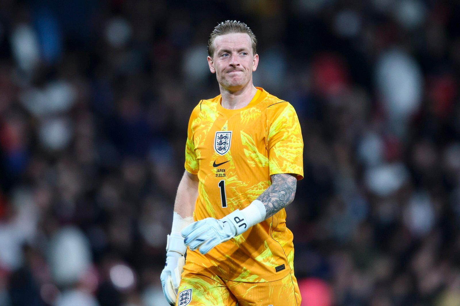 England soccer goalkeeper in orange kit with gloves, standing on the pitch as a blurred crowd watches in the background