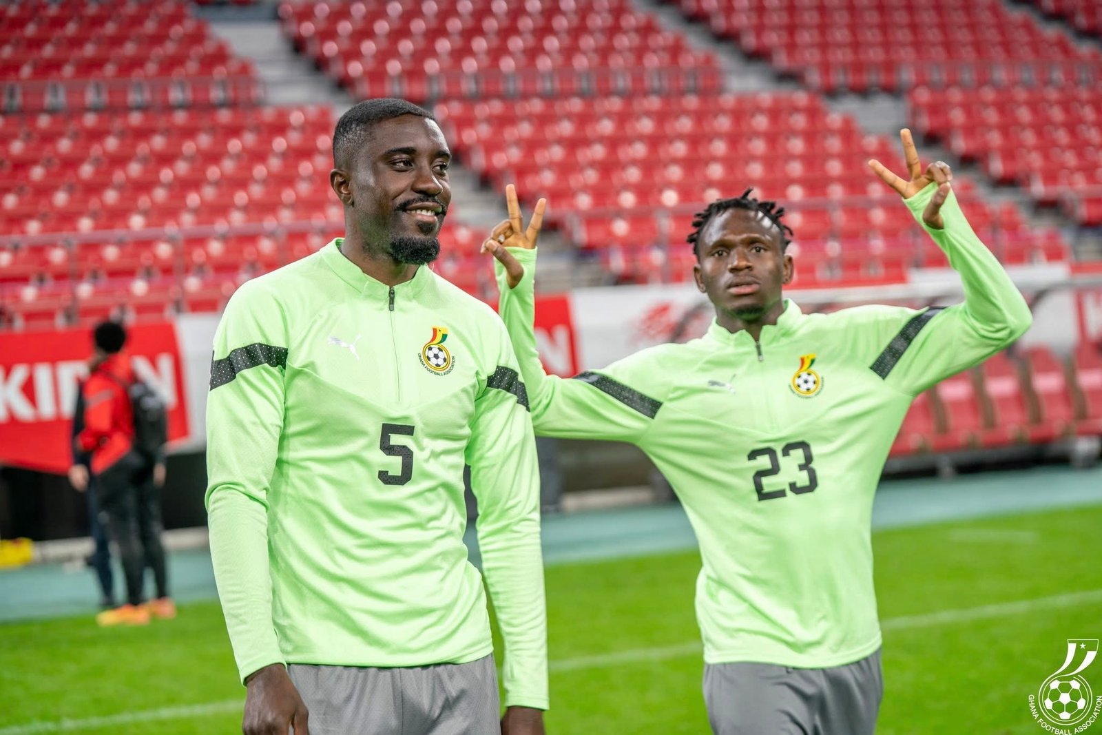 Two Ghanaian football players in mint green training tops wave with peace signs on a stadium field, red empty seats behind them. Two Ghanaian football players in mint green training tops wave with peace signs on a stadium field, red empty seats behind them.
