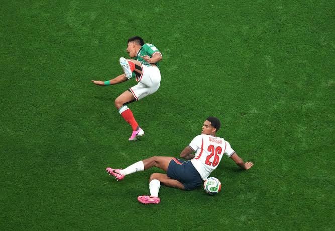 Soccer players contesting the ball on a green field; a green kit player leaps as a white kit opponent slides in. Soccer players contesting the ball on a green field; a green kit player leaps as a white kit opponent slides in.