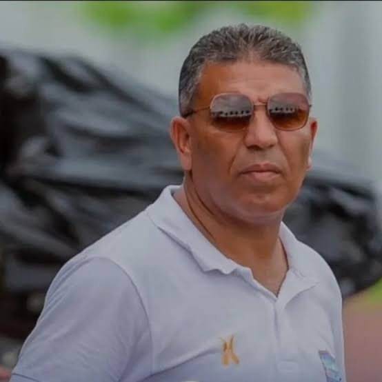 Man with short gray hair wearing sunglasses and a white polo shirt, looking to the side outdoors.