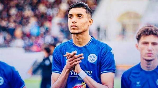Young male soccer player in a blue jersey standing on the field with hands clasped, looking ahead. Young male soccer player in a blue jersey standing on the field with hands clasped, looking ahead.