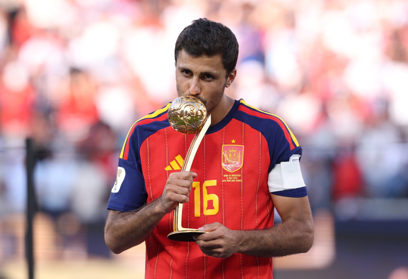 Footballer in a red Spain jersey kissing a golden trophy on a ceremony stage, stadium blurred in the background.