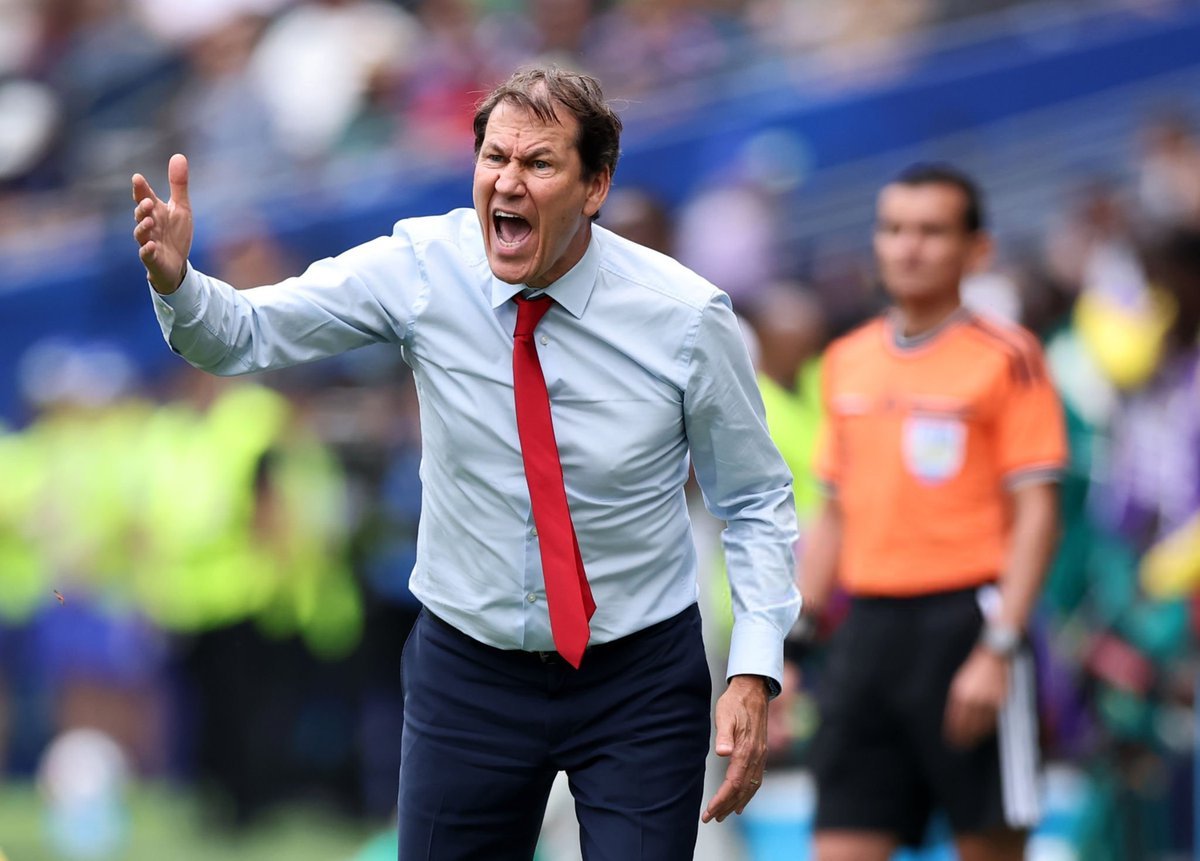 Coach on the sideline yelling angrily, wearing a light blue shirt and red tie, gesturing with an outstretched arm on a busy field.