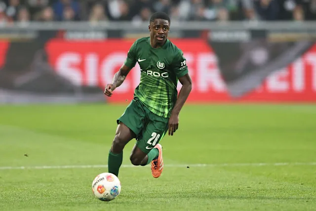 Soccer player in a green kit dribbles a ball on the grass during a match inside a stadium. Soccer player in a green kit dribbles a ball on the grass during a match inside a stadium.