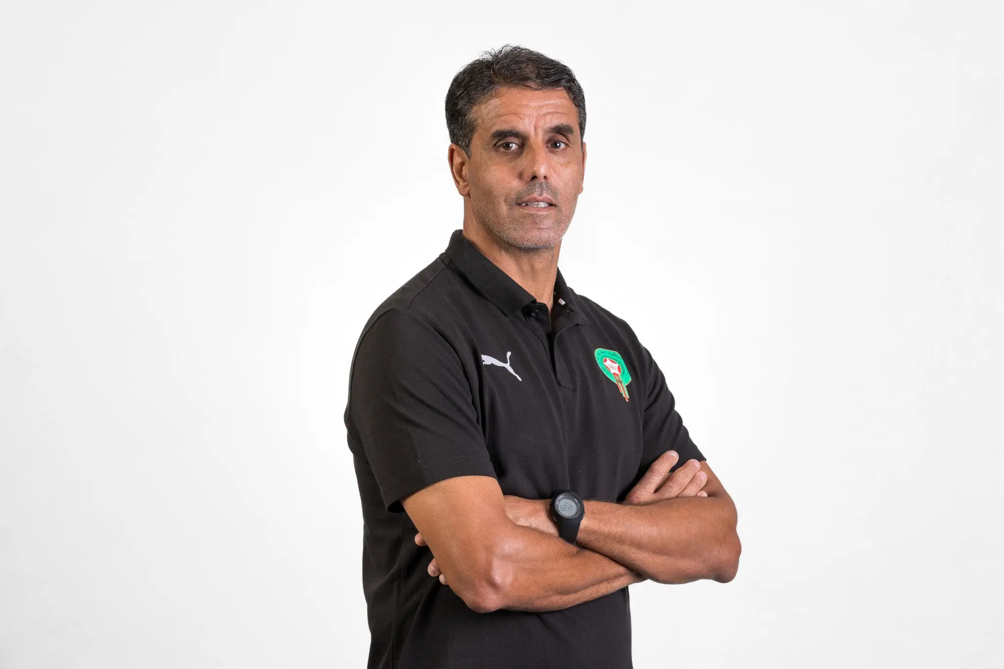 Man with short dark hair wearing a black polo, arms crossed, looking at the camera against a white background. Man with short dark hair wearing a black polo, arms crossed, looking at the camera against a white background.