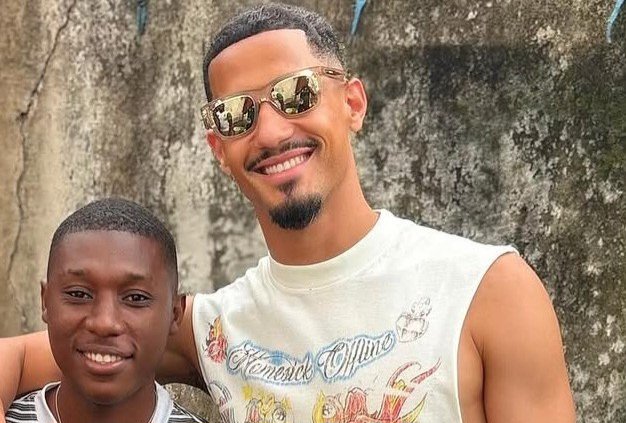 Two men smiling at the camera outdoors, with the older man wearing sunglasses and a white sleeveless graphic shirt, his arm around a younger friend.