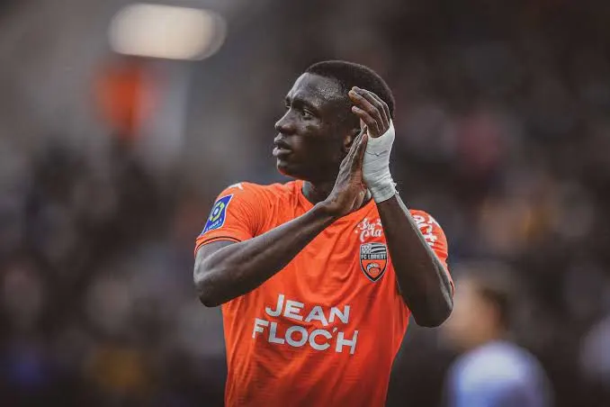 Footballer in an orange jersey clapping with hands near his face on a blurred stadium background, front reads 'JEAN FLOCH'. Footballer in an orange jersey clapping with hands near his face on a blurred stadium background, front reads 'JEAN FLOCH'.