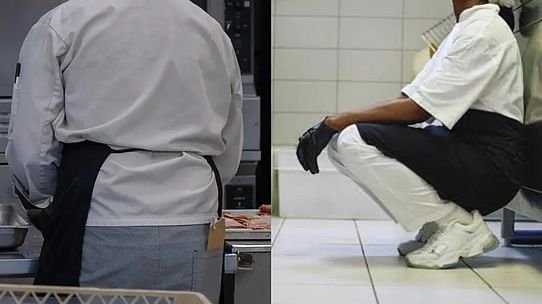 Two kitchen workers seen from behind: a standing chef at a prep counter on the left and a crouching worker in white clothes on the right. Two kitchen workers seen from behind: a standing chef at a prep counter on the left and a crouching worker in white clothes on the right.