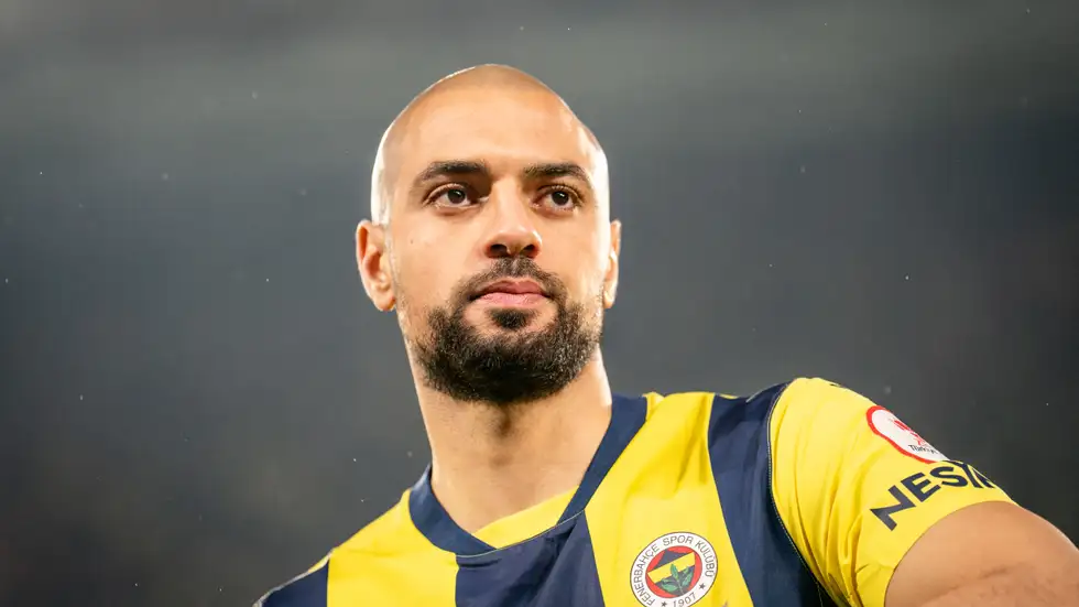 Male soccer player with a shaved head and beard, wearing a yellow and navy jersey, looking pensively into the distance on the field.