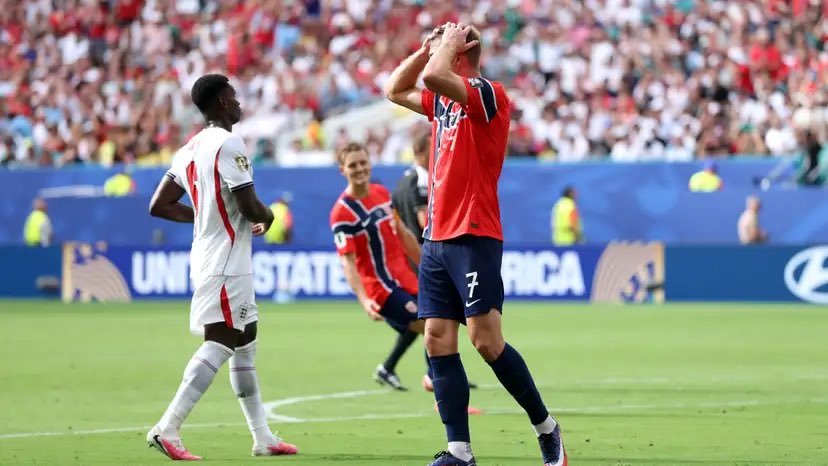 Soccer players stand on a green pitch as a player in red holds his head in frustration while a white-jersey opponent looks on, with a packed stadium in the background. Soccer players stand on a green pitch as a player in red holds his head in frustration while a white-jersey opponent looks on, with a packed stadium in the background.