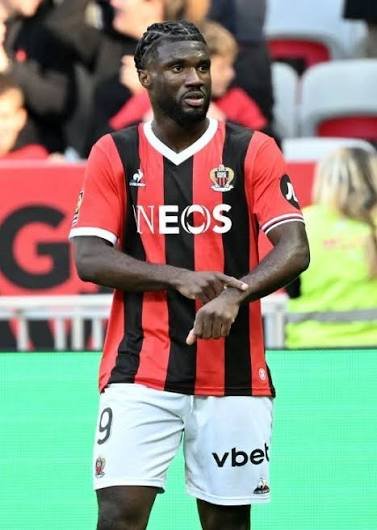 Soccer player in red and black striped kit with INEOS sponsor, standing on the field with arms crossed; number 9 on white shorts.