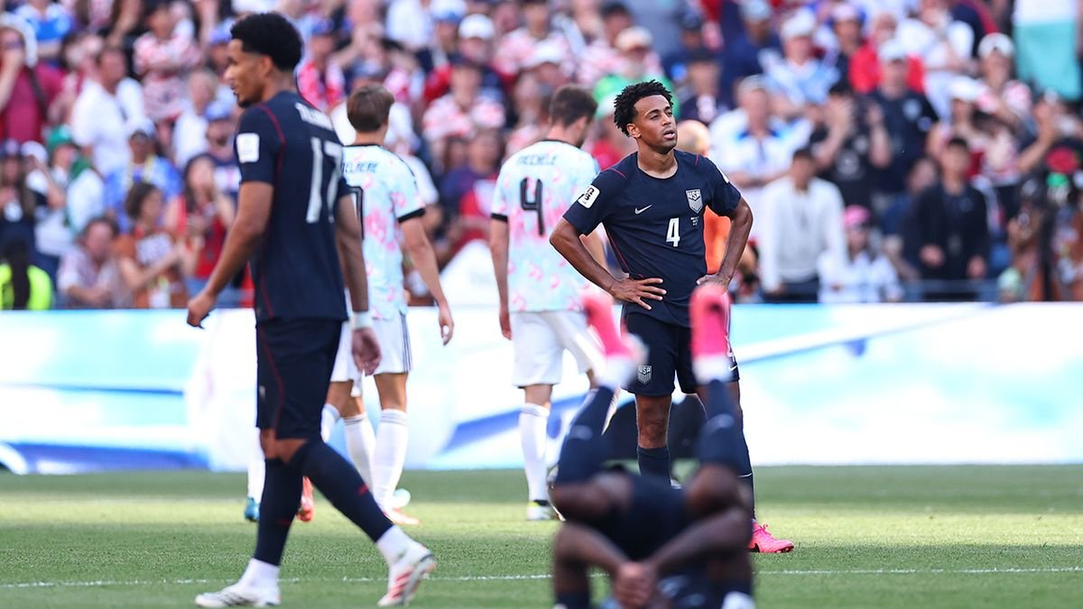 USA soccer player wearing number 4 stands with hands on hips on the field as teammates walk by and a large crowd watches in the stands.
