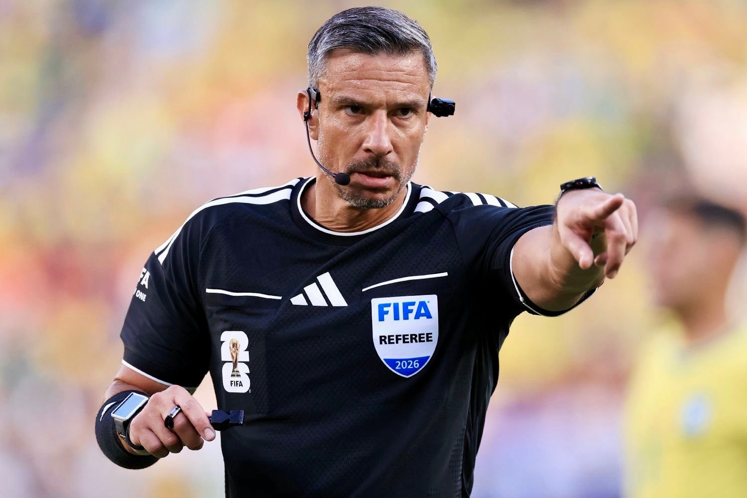 FIFA referee in black shirt, headset, and wrist devices points decisively during a match with a colorful crowd in the background.