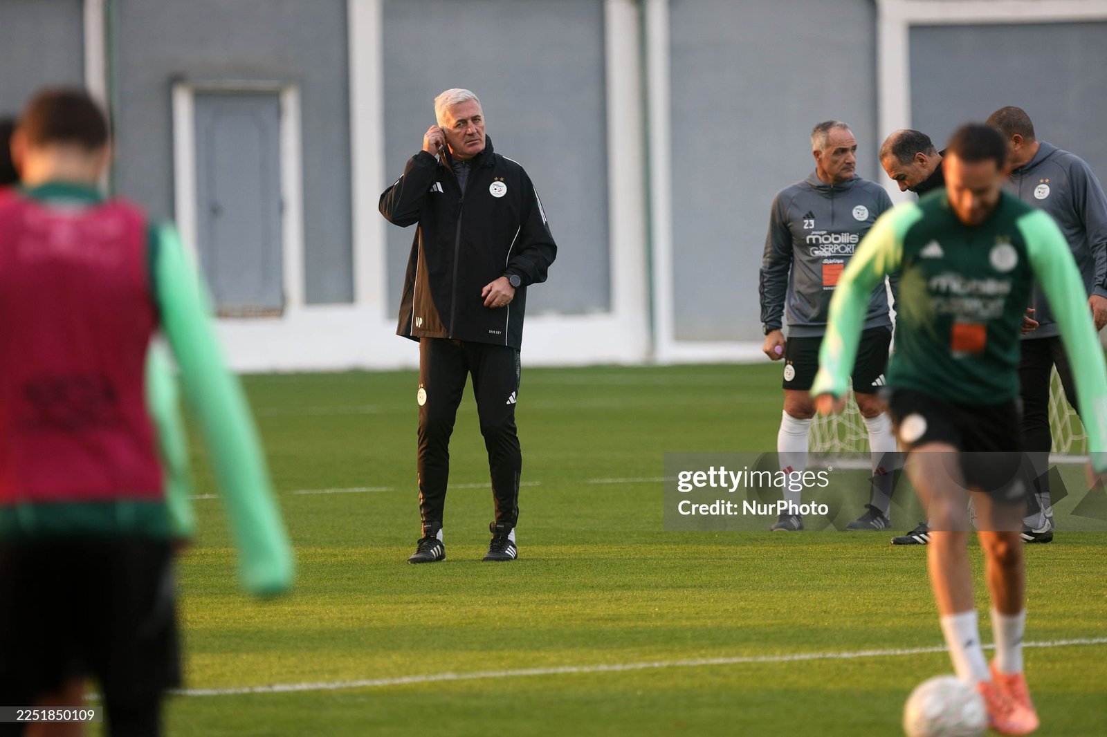 Coach in a black tracksuit on a soccer field, talking on a mobile phone during training with players practicing in the background.