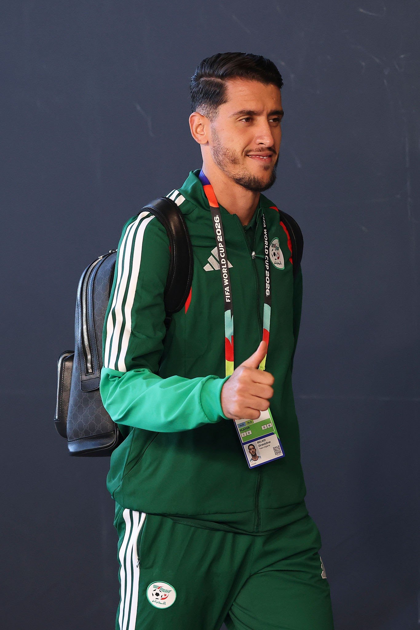Man in a green athletic tracksuit with a backpack and event lanyard, standing against a dark wall.