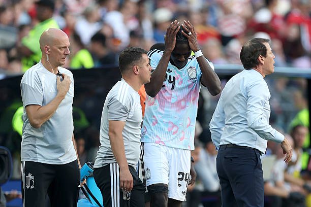 Soccer players and a coach on the sideline; the central player holds his head in distress while teammates look on. Soccer players and a coach on the sideline; the central player holds his head in distress while teammates look on.