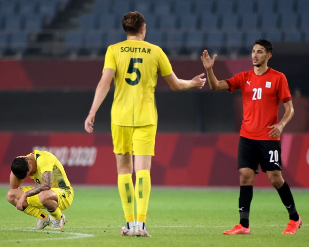 Soccer players shake hands on the field after a play: yellow team number 5 faces away, red team number 20 reaches out, and a yellow teammate sits on the grass nearby.