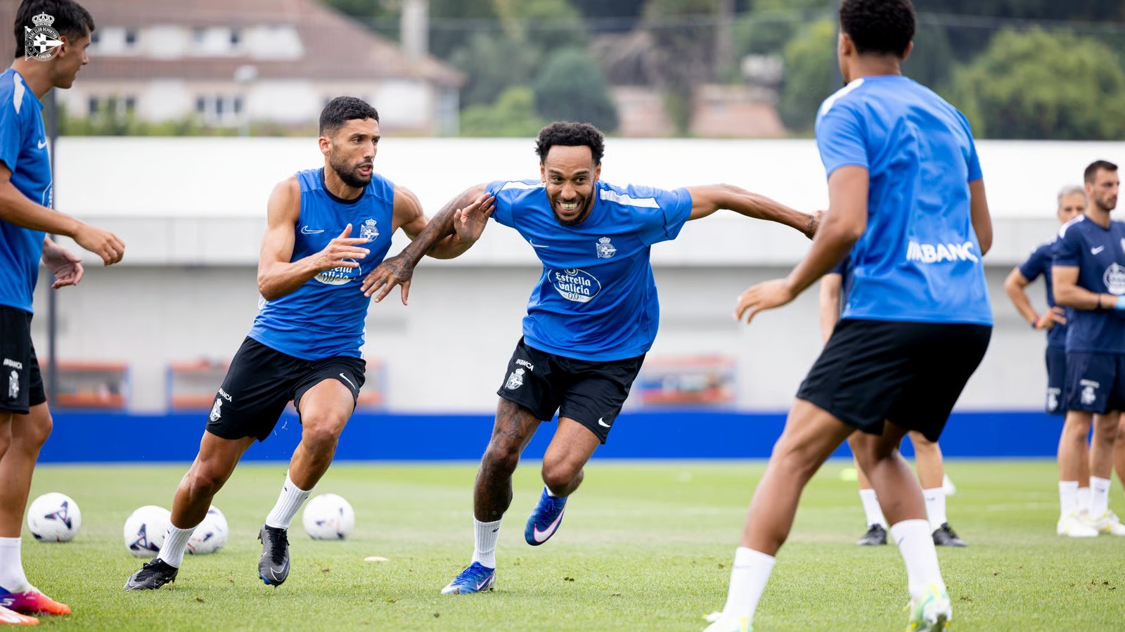 Soccer players in blue jerseys train on a green field, chasing balls during a practice drill, with a team crest visible on their shirts.