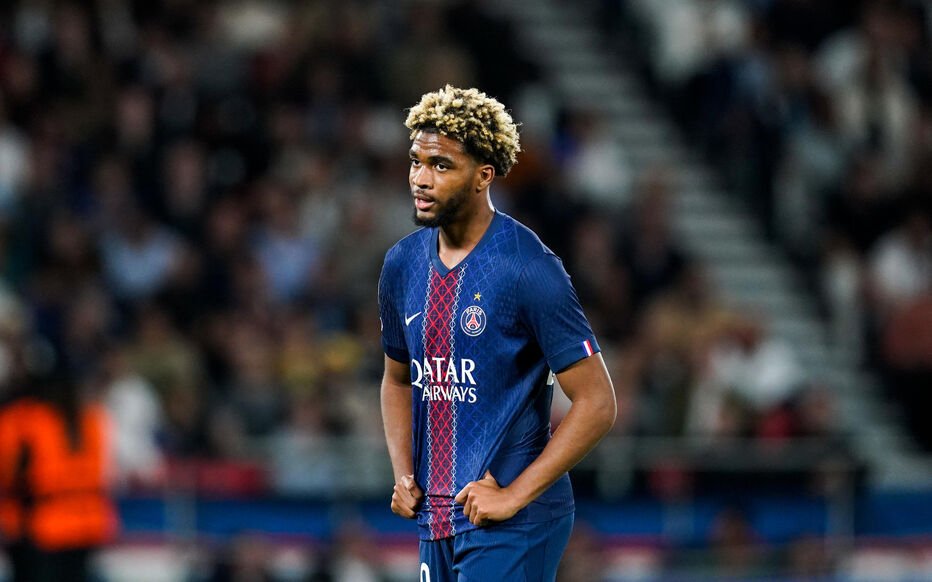 Soccer player in a blue Paris Saint-Germain jersey stands with hands on hips on the field, crowd in the background.