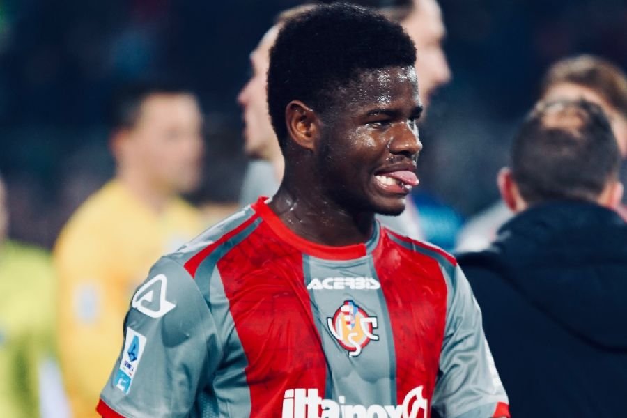 Soccer player in a red and gray jersey celebrating on the field, tongue out and smiling.