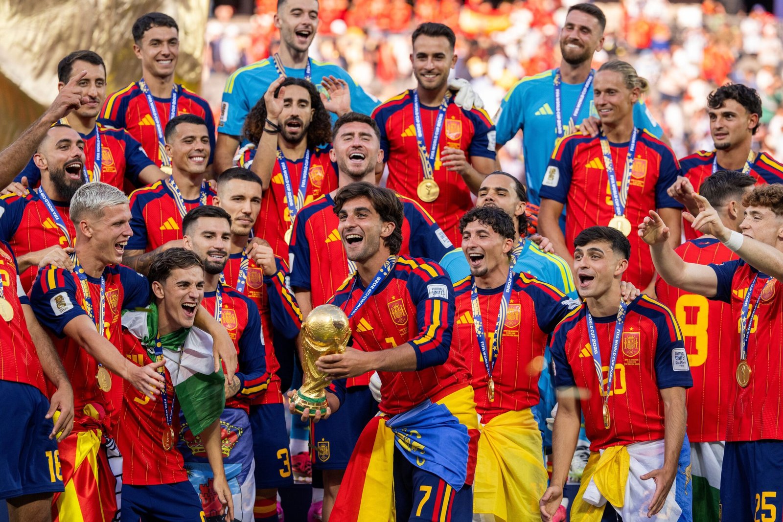 Spain national football team players celebrate a victory with medals and a gold trophy in a crowded stadium crowd behind them, wearing red and blue striped jerseys