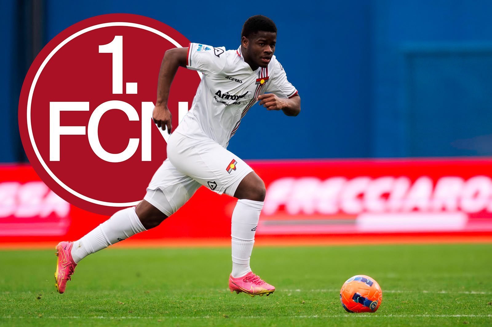 Soccer player in a white kit sprinting after an orange ball on a green field, with a large red 1. FCN logo in the background badge.