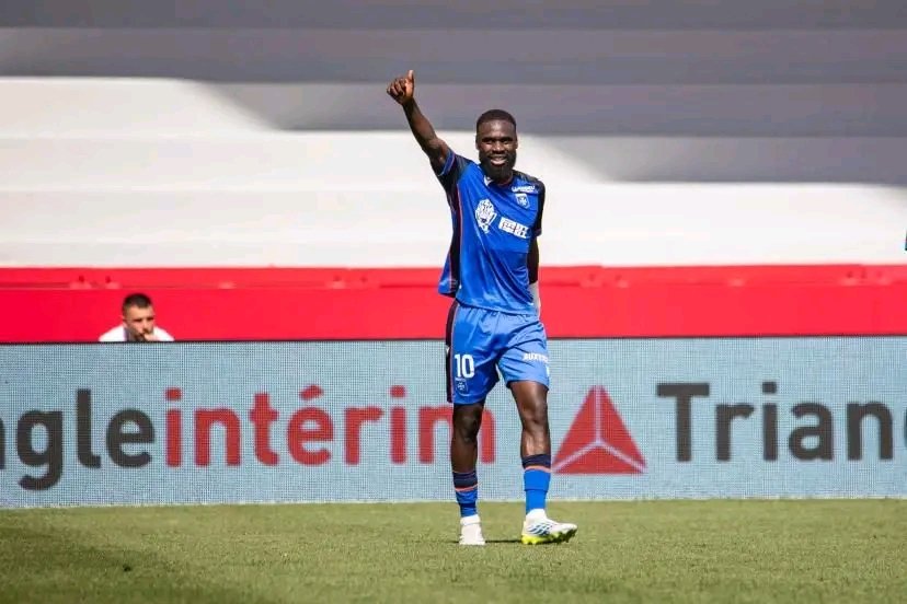 Soccer player in blue kit with number 10 raising a fist in celebration on the pitch, with ads in the background.
