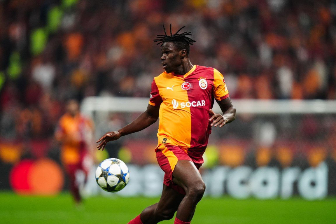 Soccer player in orange-and-red Galatasaray jersey chasing a ball during a match, stadium crowd in the background.