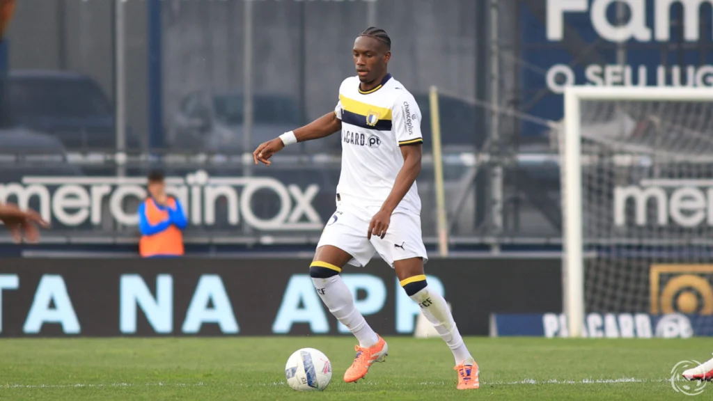 Soccer player in a white jersey with yellow and navy accents dribbles an orange cleat ball on a grassy field, in action during a match.
