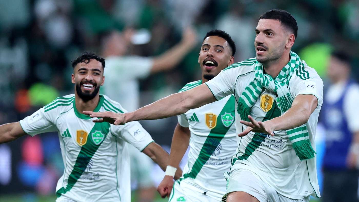 Three soccer players in white and green jerseys celebrate on the field, smiling as they run forward together after a goal or win, with a green scarf visible on one player in the foreground.
