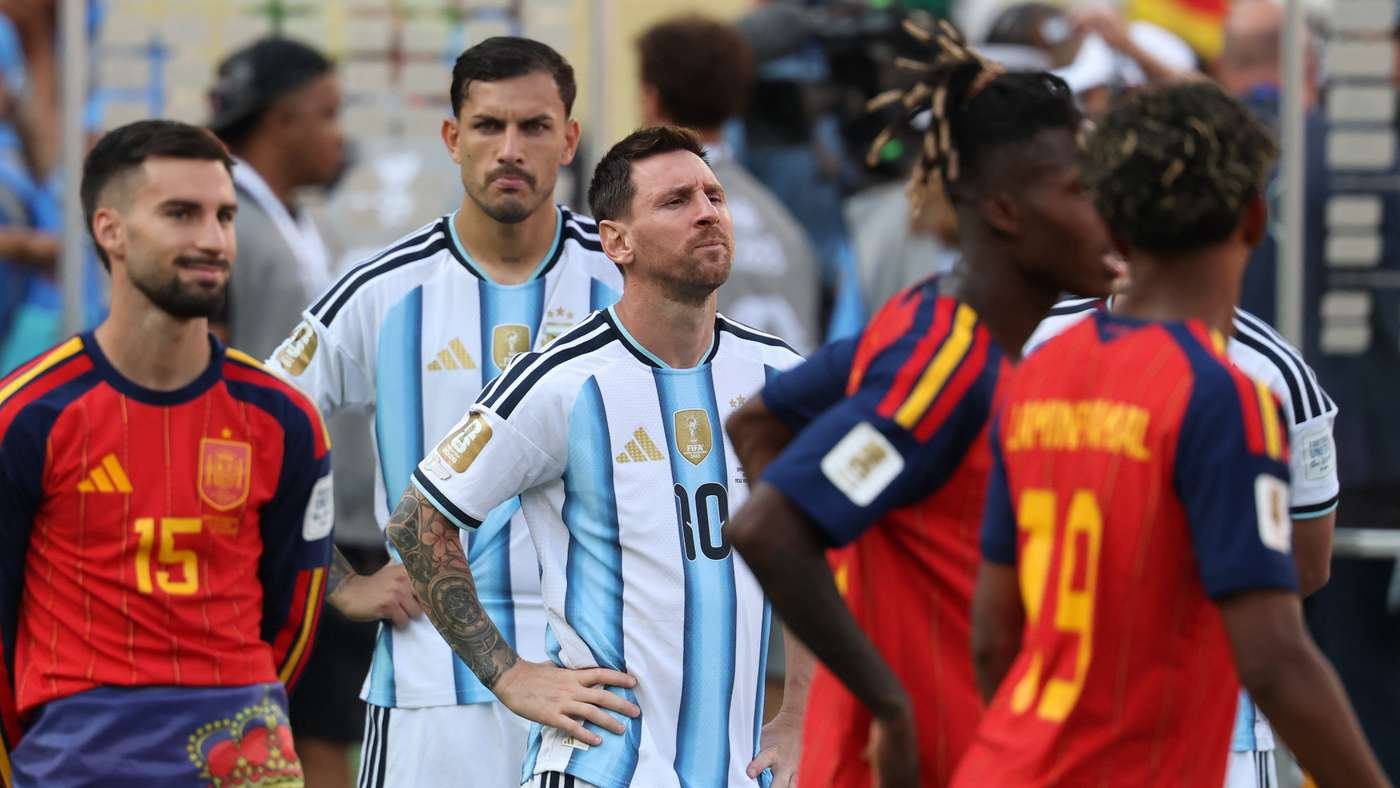 Argentina players in light blue and white stripes stand with Spanish players in red jerseys on the field before a match.