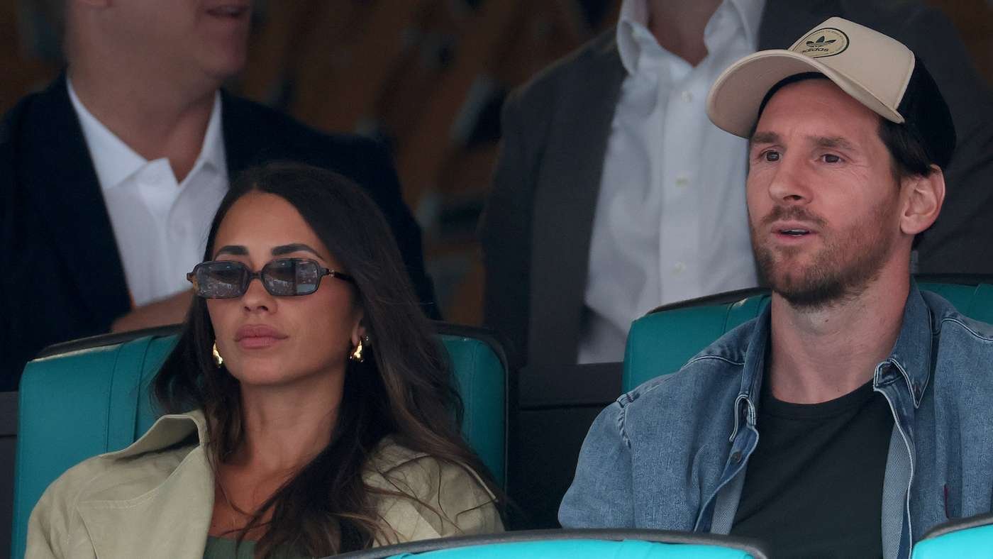 A man in a denim jacket and cap sits beside a woman wearing sunglasses in turquoise stadium seats, watching an event.