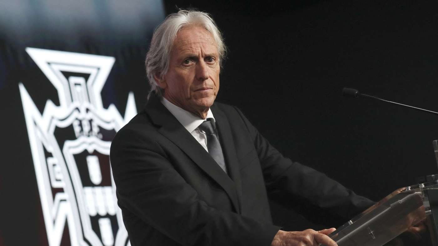 Older man in a black suit and tie speaking at a podium on a stage, with a large crest emblem in the background.