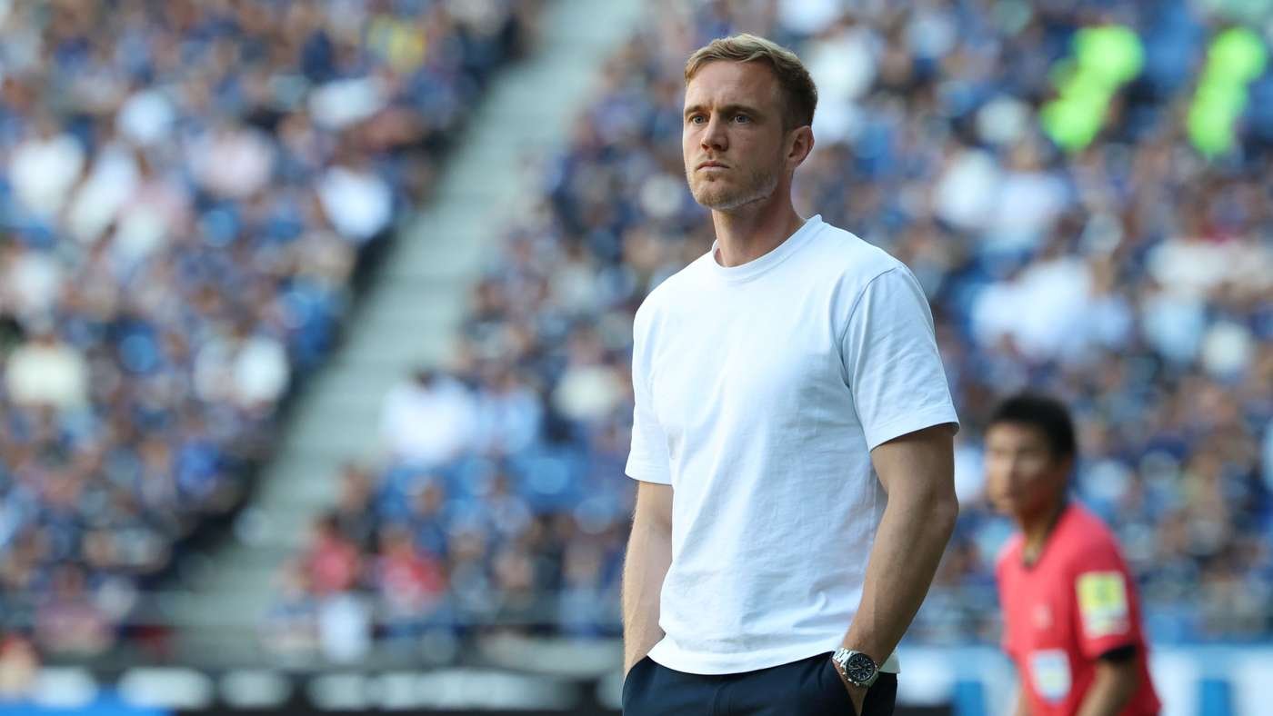 Man in a white T-shirt standing on a stadium field with a blurred crowd in the background.