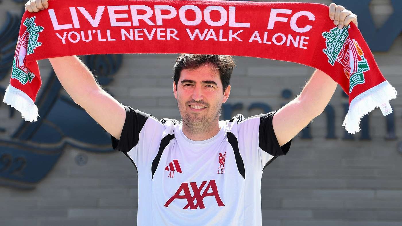 Man in a white Liverpool FC jersey holds up a red scarf reading 'Liverpool FC You'll Never Walk Alone'
