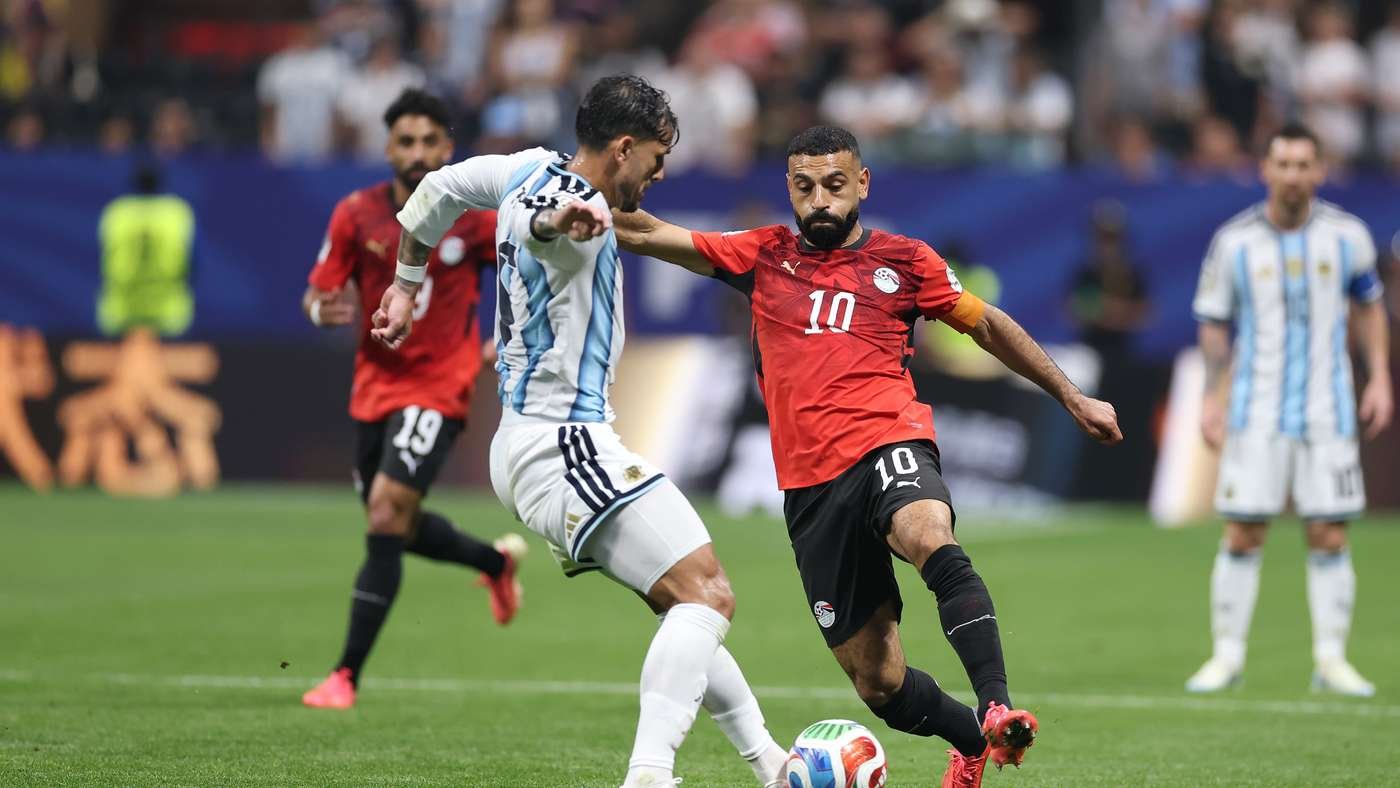 Soccer players chase the ball during a match; one in light blue and white stripes, the other in red with number 10 and an armband.
