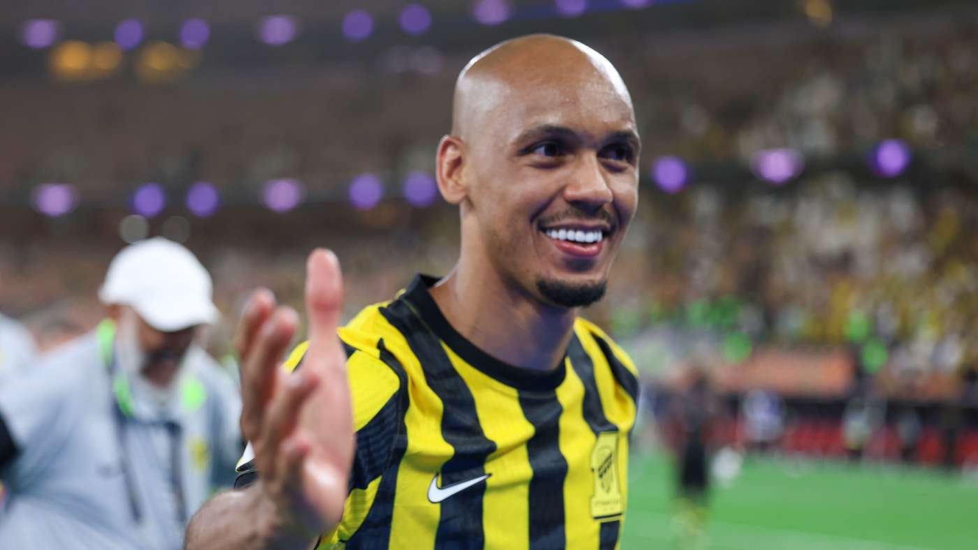 Soccer player in a yellow and black striped jersey smiles and extends a hand in front of a blurred stadium crowd.