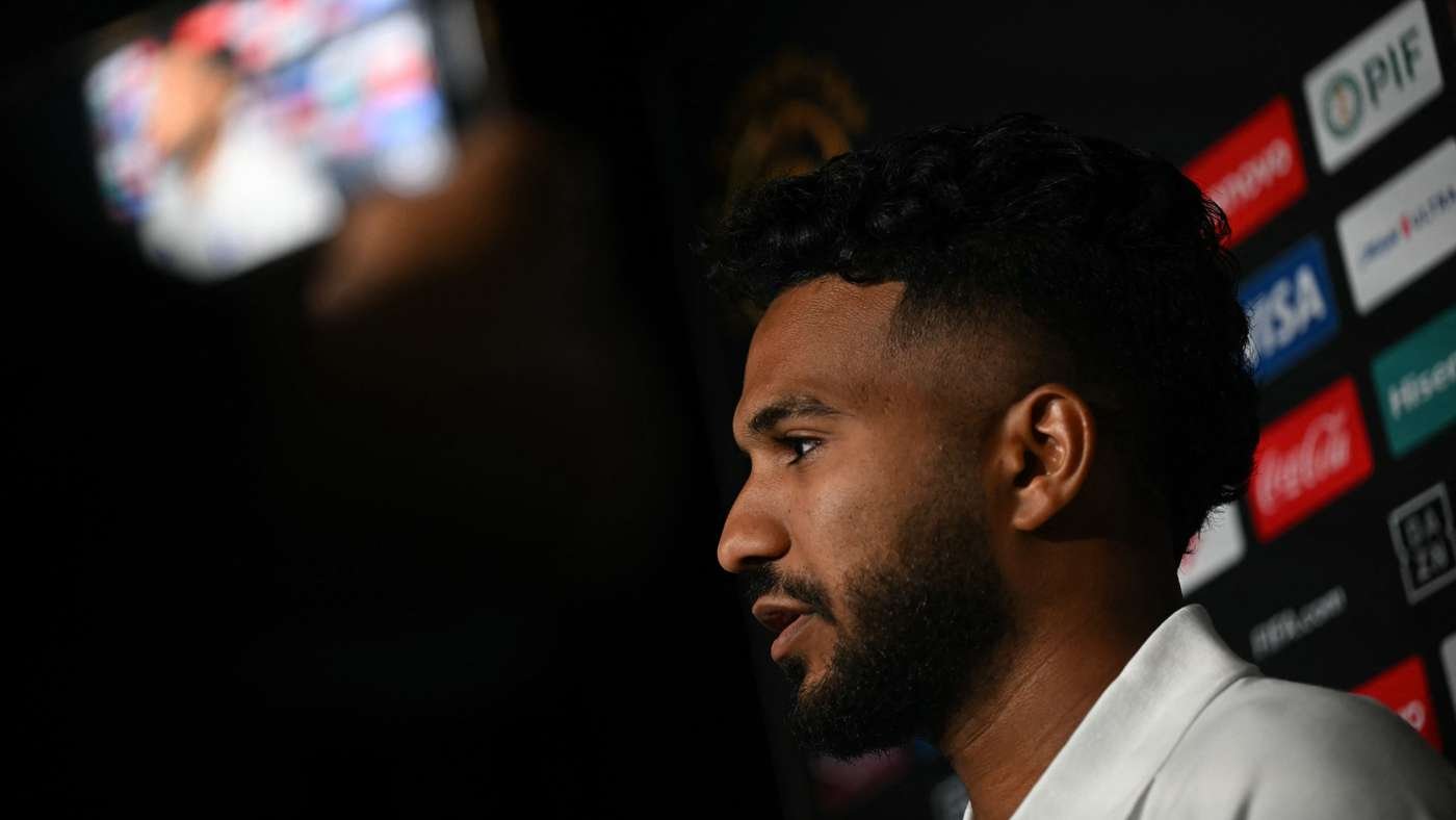 Profile view of a bearded man speaking at a press event, with sponsor logos in the backdrop.