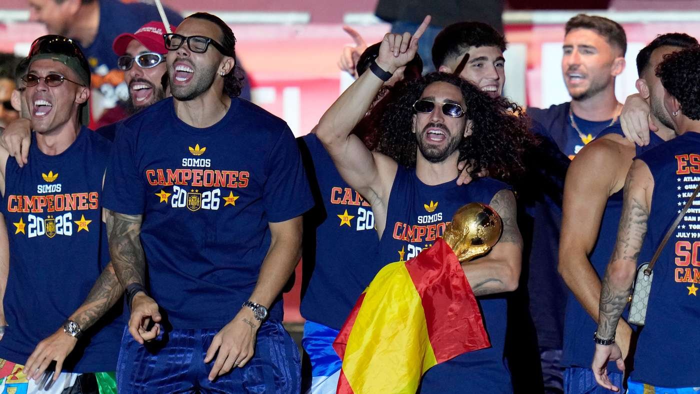 Group of male athletes celebrating a championship, wearing blue shirts that read 'Somos Campeones 2026', with a gold trophy and a Spanish flag visible.