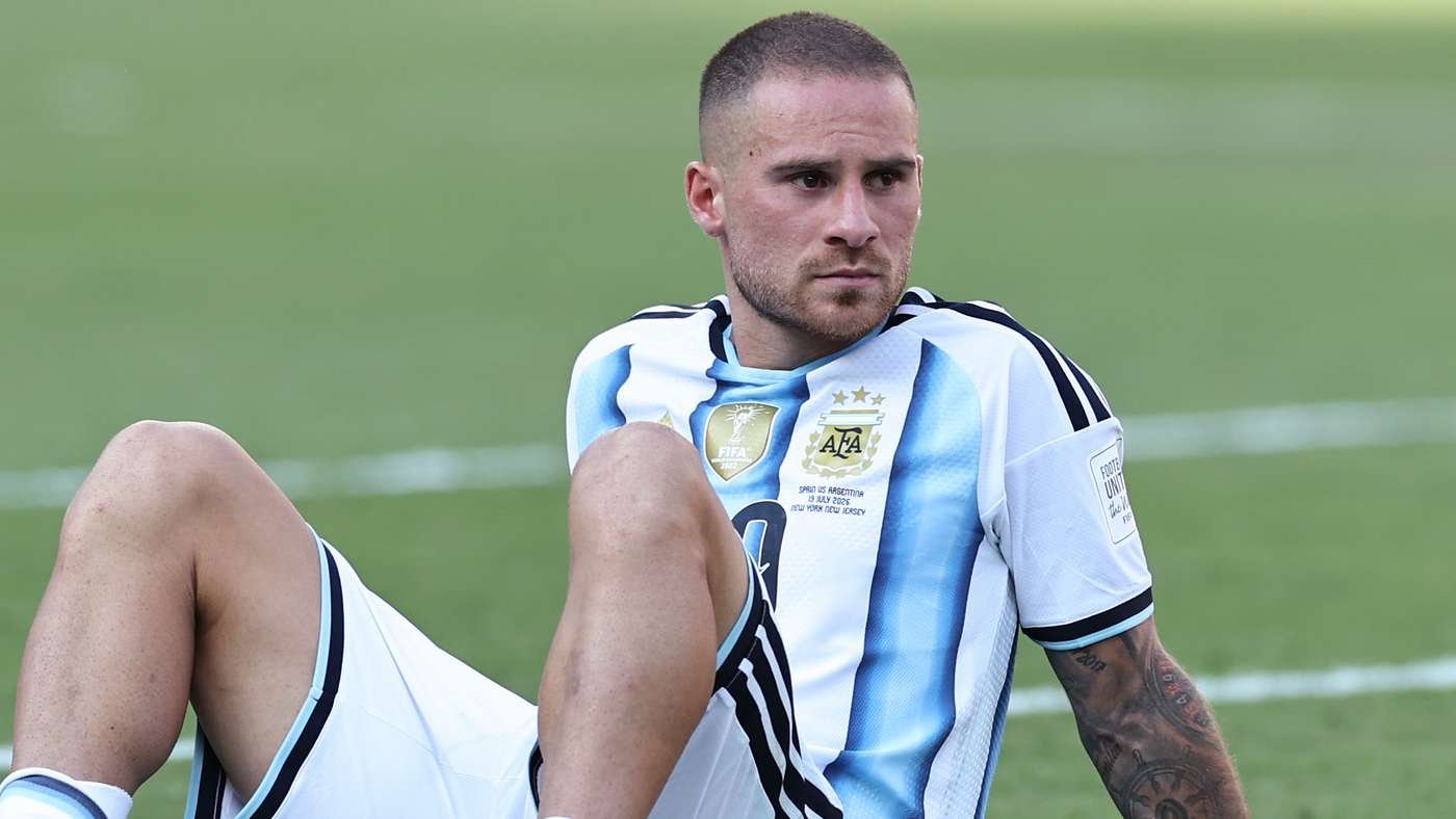Soccer player in Argentina-style jersey sits on a grassy field, looking off to the side.