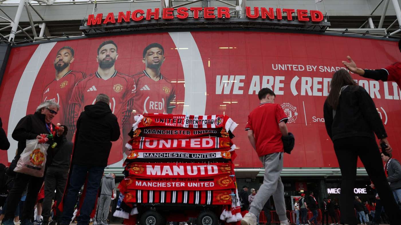 Group of fans in red outside Manchester United stadium, with a large red mural of players and stacked Manchester United scarves in the foreground.
