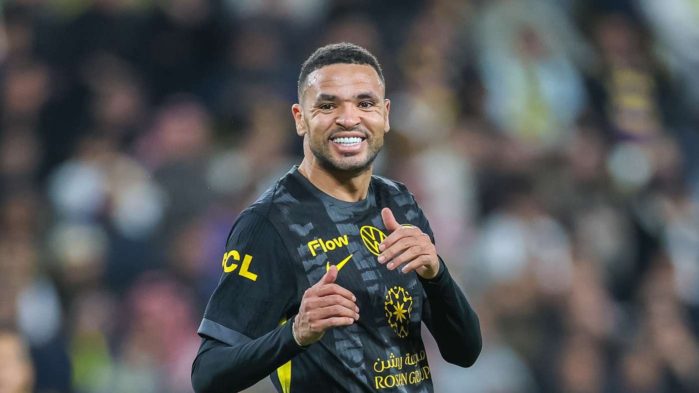 Smiling male soccer player in a dark jersey with yellow logos, stadium crowd in the background.