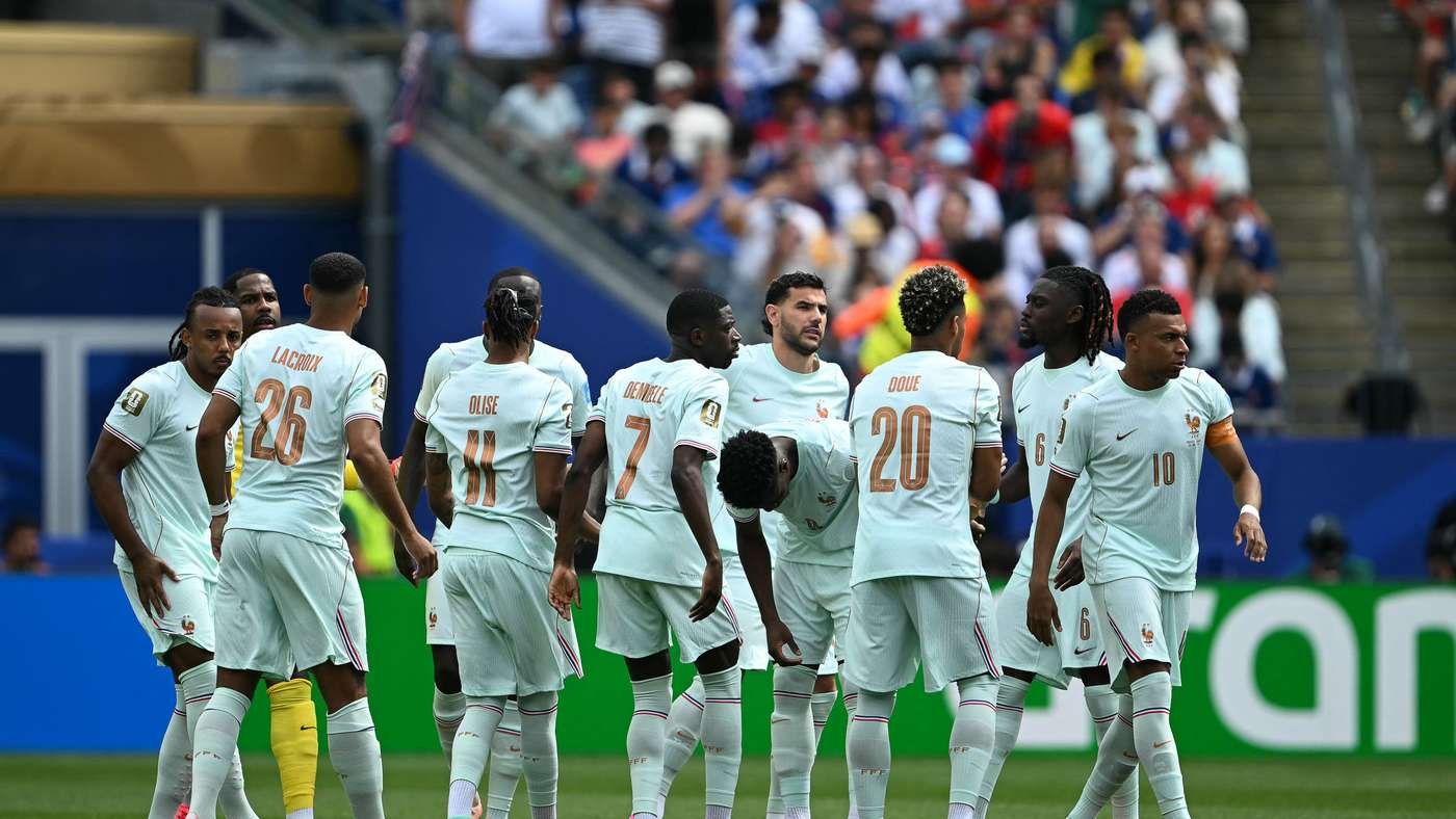 A group of France national team players in pale blue uniforms stand together on a soccer pitch, backs visible with numbers 26, 11, 7, 20, amid a crowded stadium fans in the background.