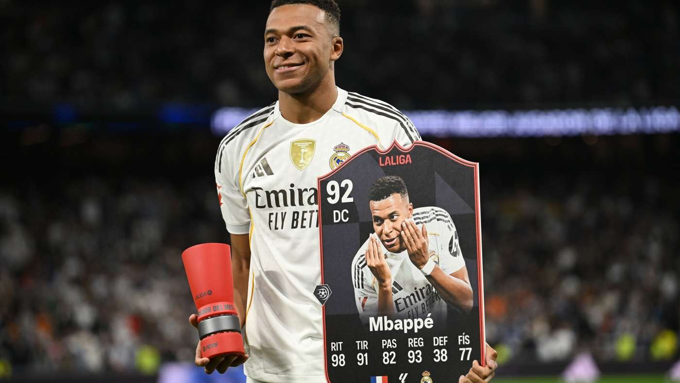 Soccer player in a white Real Madrid jersey on a field, smiling, holding a large La Liga card featuring his portrait and the name Mbappé with stats at the bottom.