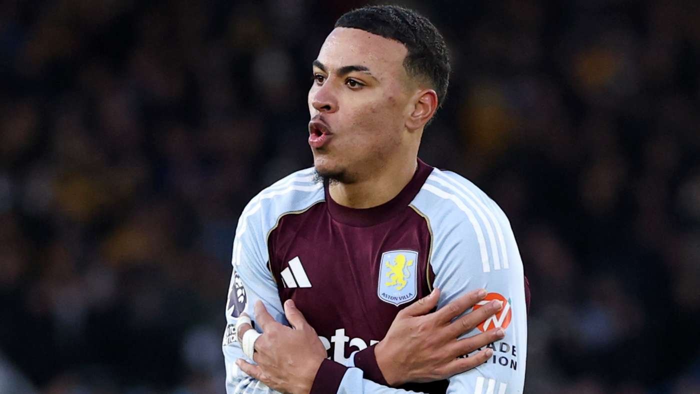 Aston Villa footballer in maroon and light-blue kit, arms crossed over his chest during a match celebration gesture .