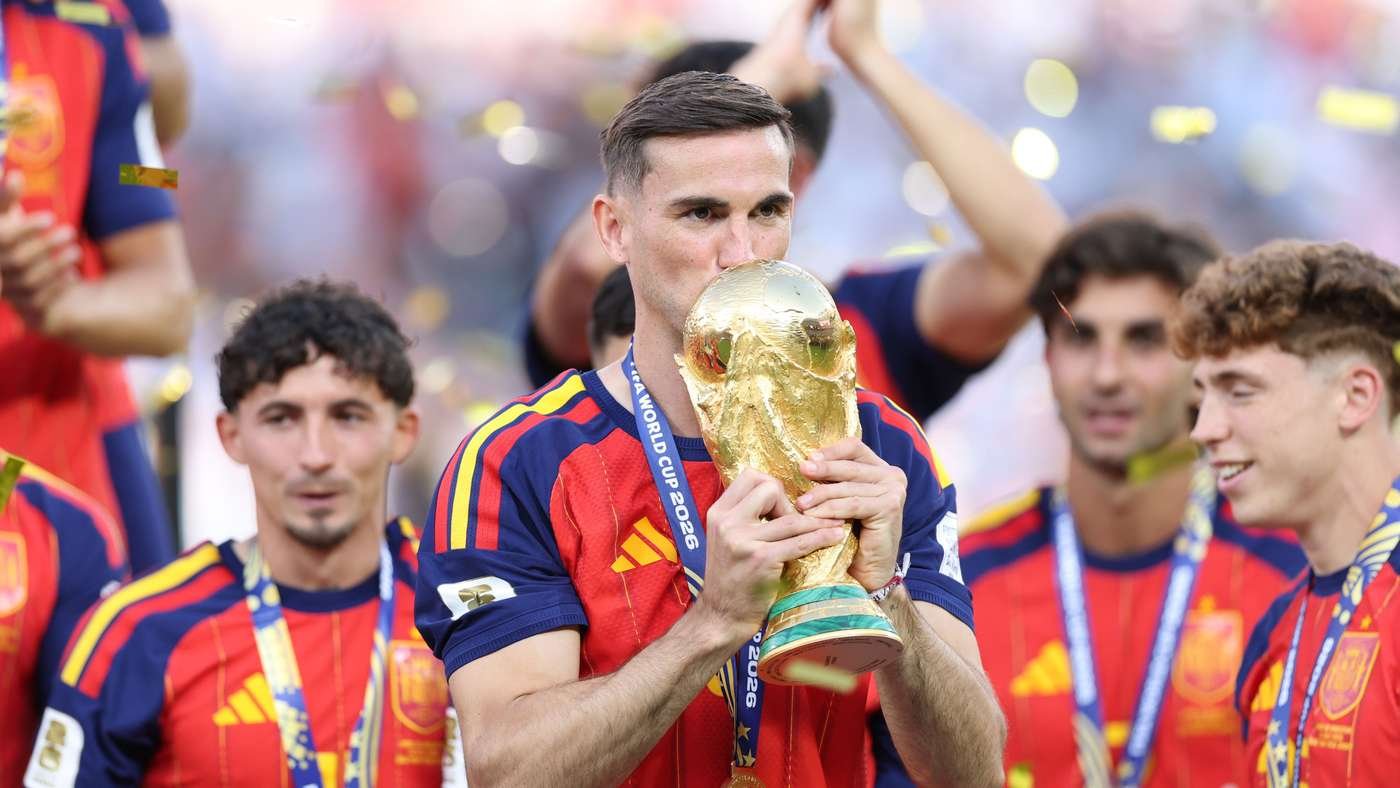 Soccer player kisses a gold trophy while teammates celebrate with medals and confetti behind him