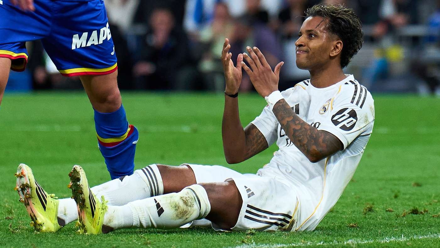 Soccer player in white sitting on the grass, clapping while a blue-clad opponent stands nearby during a match. Soccer player in white sitting on the grass, clapping while a blue-clad opponent stands nearby during a match.