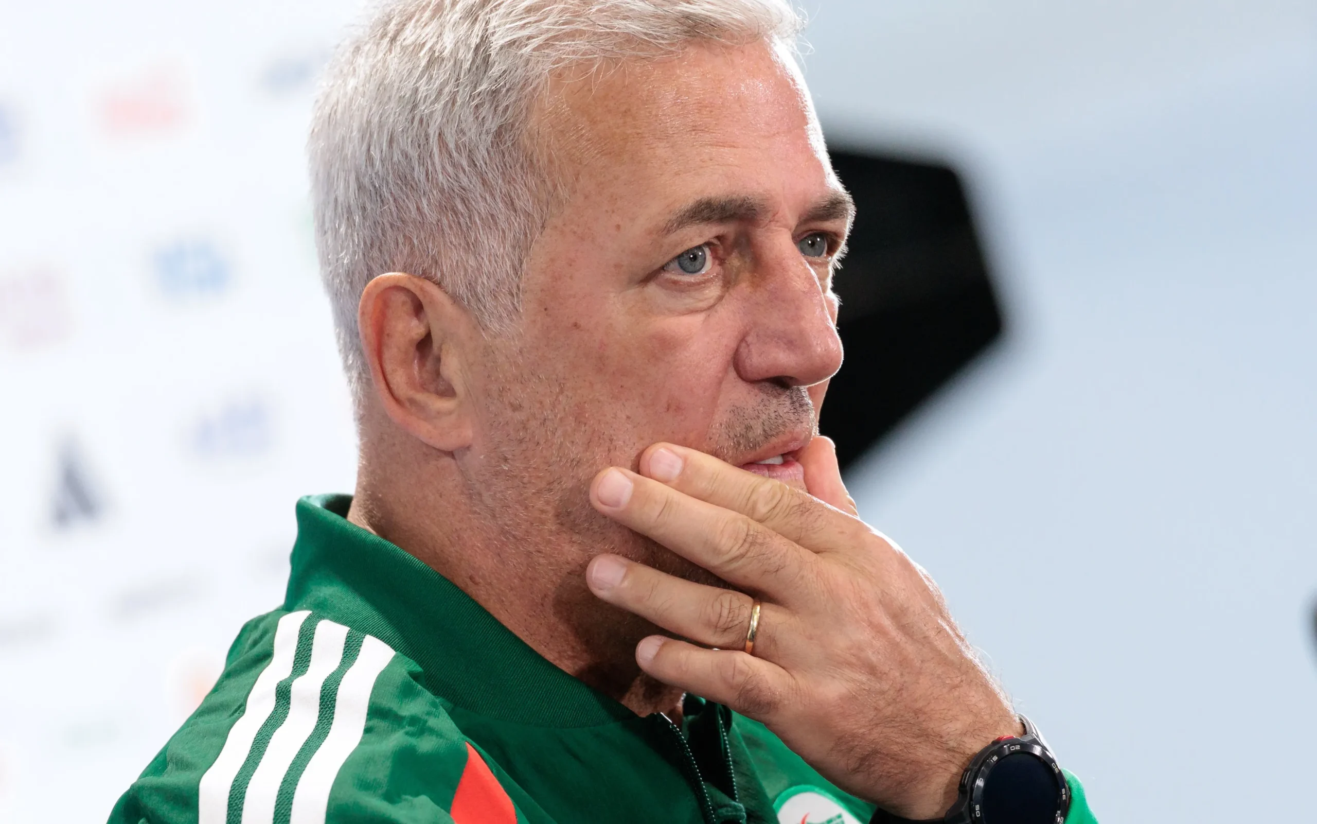 Man with gray hair in a green athletic jacket touching his chin in a thoughtful pose (side profile).