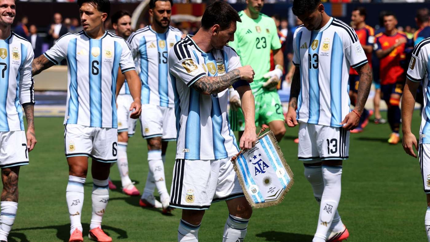 Argentine national team on the field, one player adjusting his sleeve while others stand nearby, holding a flag with the AFA emblem. Argentine national team on the field, one player adjusting his sleeve while others stand nearby, holding a flag with the AFA emblem.