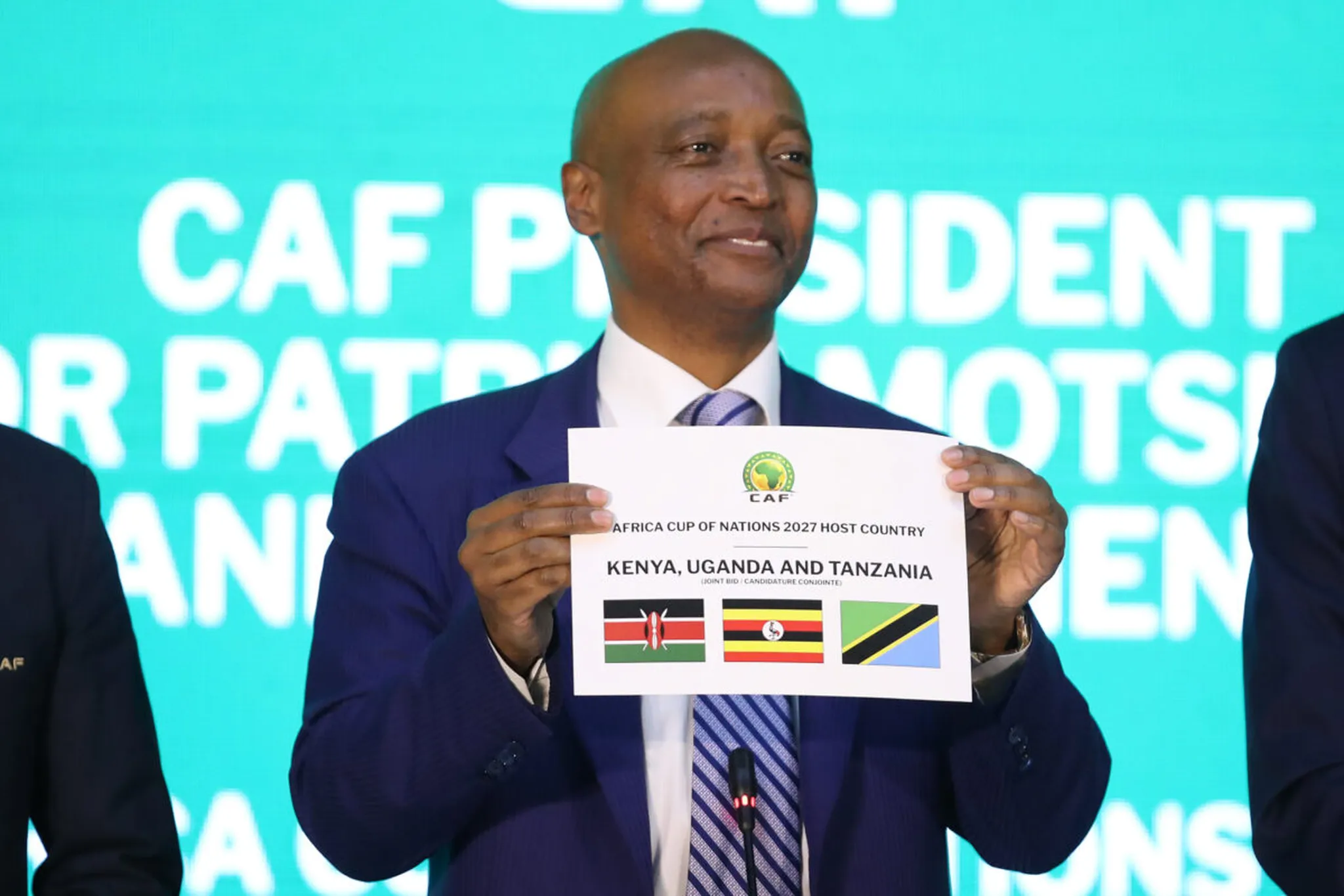 Man in a dark suit holds up a flyer with AFCON 2027 hosts Kenya, Uganda and Tanzania, and three national flags in front of a bright turquoise backdrop. Man in a dark suit holds up a flyer with AFCON 2027 hosts Kenya, Uganda and Tanzania, and three national flags in front of a bright turquoise backdrop.